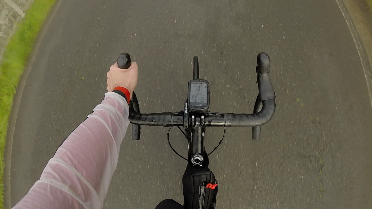 A picture taken from my point of view while riding the bike, looking down, my left hand is on the handlebar and the garmin is showing km 186.2. The background is a narrow road with its asphalt all wet