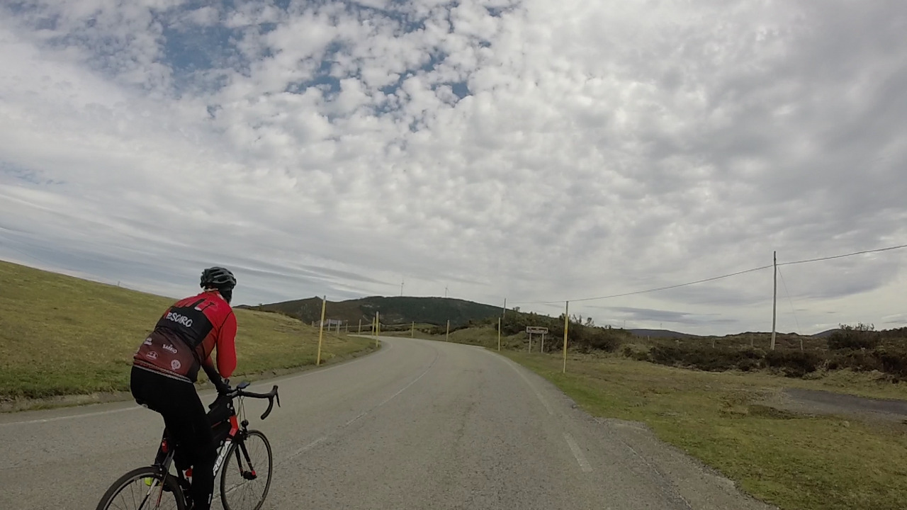 The last turn, at the top of a Garganta. Dani is on the left of the picture, riding towards it. On the left of the turn, far in the photo, a tiny sign says 'A Garganta - 900m'. The sky is full of small clouds, resembling a cobblestones road.