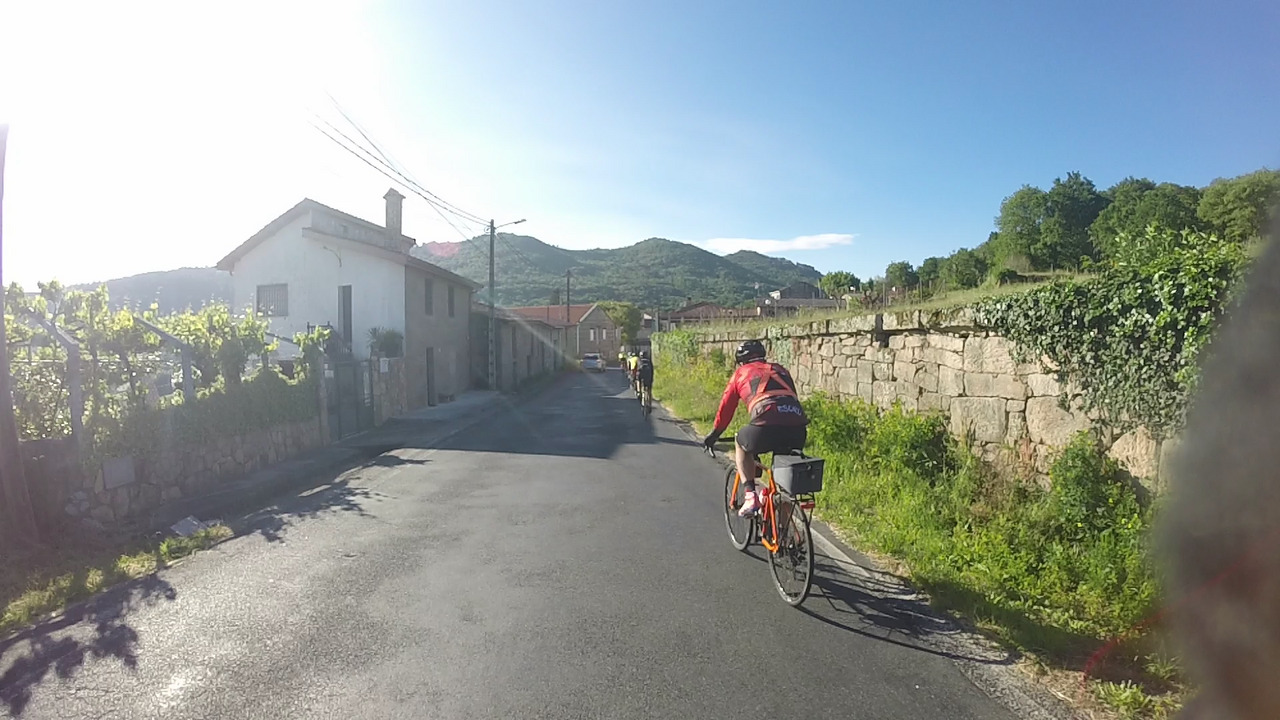 Edgar, riding on a small road, passing a house that has some land with vines on the left, and a stone wall on the right. The sky is blue and cloudless
