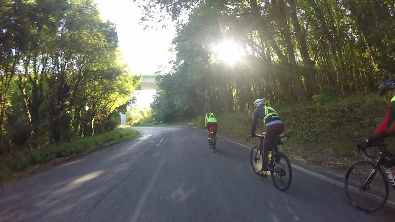 Dani, Alex and Jose riding towards the sun, the road turns a bit to the left and goes under a bridge