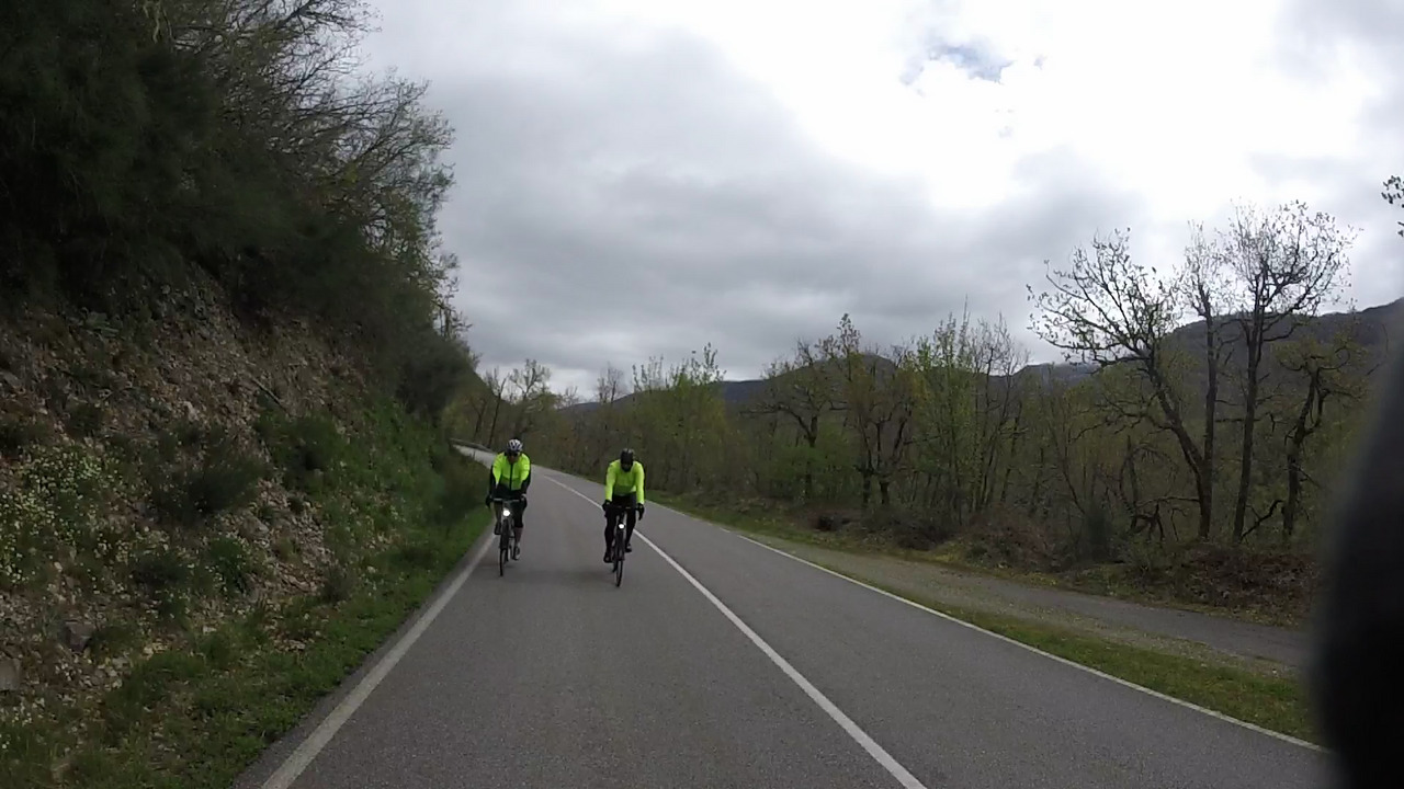 Ferreiro and Dani, chasing me down the road. A side of a hill is on the left of the photo, while the right of the photo show some naked trees and the mountains surrounding the valley we were in