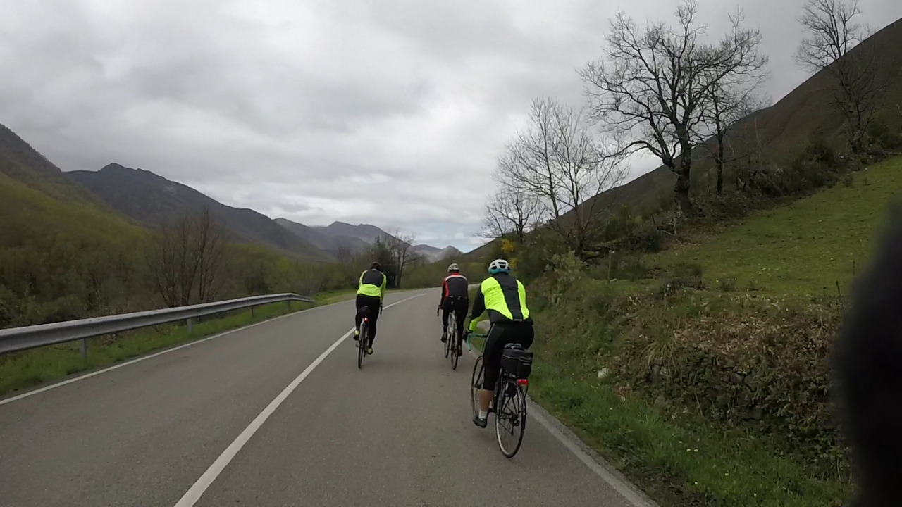 Dani, Juankar and Ferreiro riding on a road that turns slightly right, there is a hill covered with some trees to their right, an open view of the mountains to their left