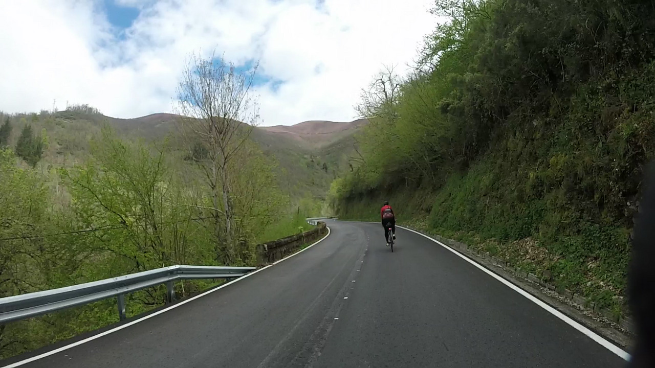 Dani riding alone on a road, going downhill with some turns. The road has the side of a hill to the right and some naked trees to the left. High mountains can be seen at the back of the photo