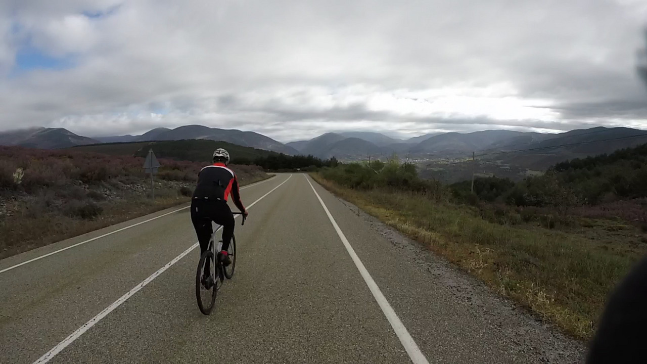 Juankar going down a straight big road. The sun is trying to shine from behind the mountains at the back of the photo, but there are too many grey clouds
