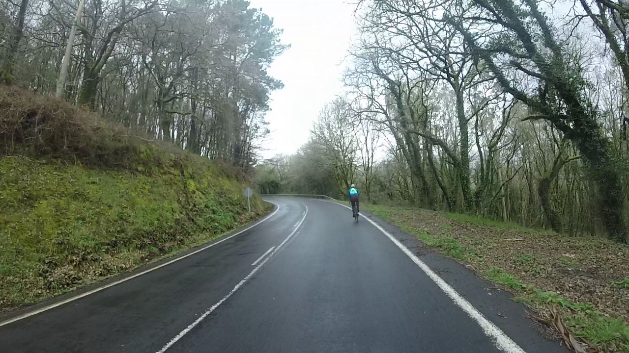 Alejandro standing on his bike, more climbing towards the 4th control point on the same wet road as before, with trees on the right and a small concrete building on the left