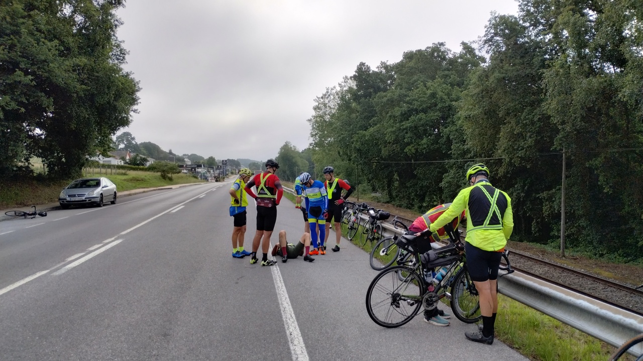 Alex, laying on the road after the crash, several randonneurs are around him, while 2 others are looking at the bike that caused the crash