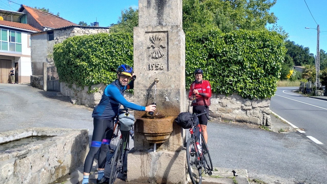 Me, refilling my water bottle at the fountain in alto do couso. Dani is behind the fountain to the right. The fountain has a drawing of a bow with 5 arrows carved on it, as well as the numbers 1956.
