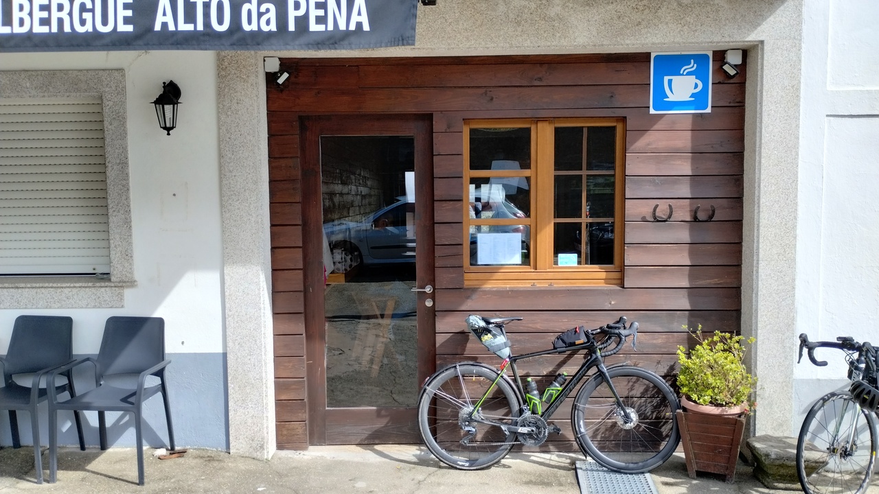 The synapse leaning by the entrance to the café/hostel. A sign on the top-right says 'Albergue Alto da Pena', the entrance is made of wood and there is a door with a big glass and a window with 4 smaller glasses by it, over my bike. A blue sign over the window has an icon of a steaming cup