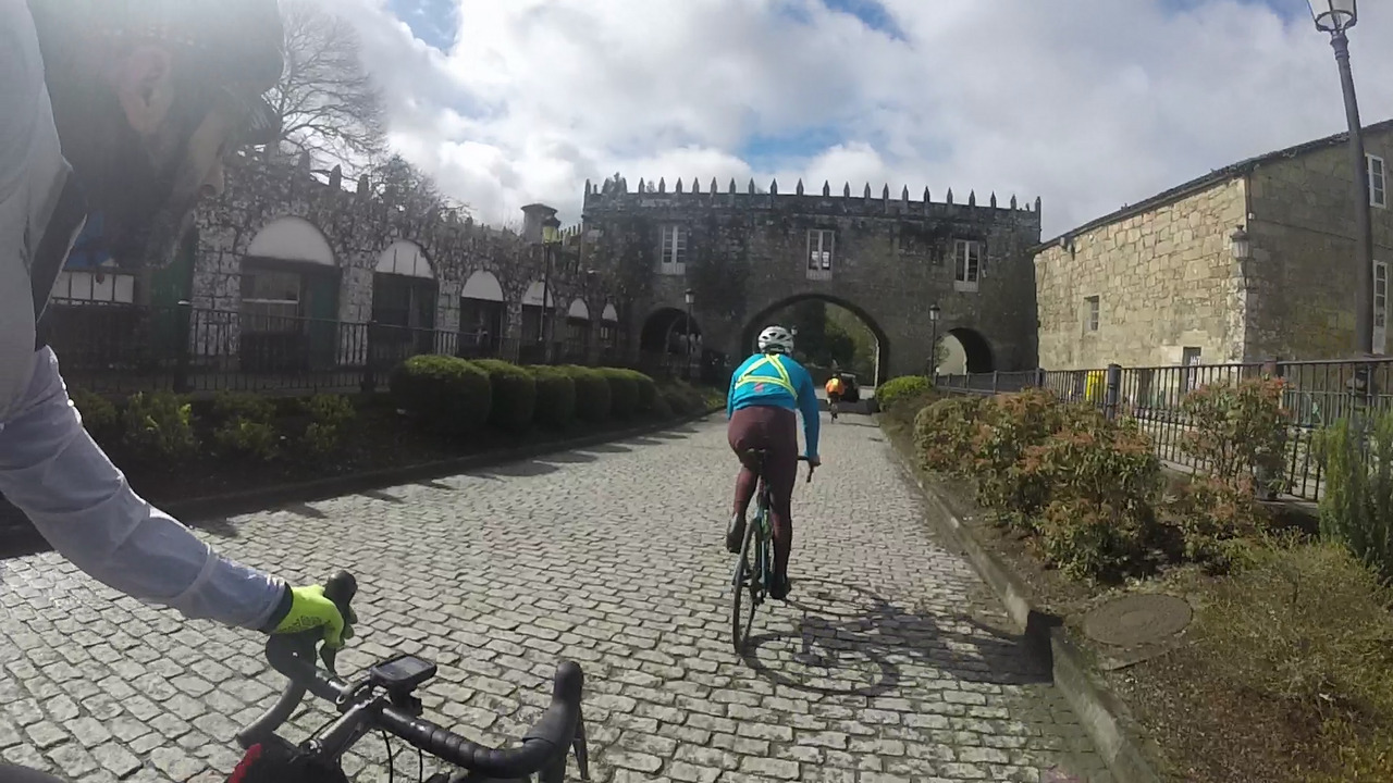 The cobblestones path that leaves Negreira, Alejandro is riding in front of me, I'm on the left side, one hand on the handlebars, the other recording with the gopro. Old structures made of stone, resembling a castle entrance, can be seen at the back
