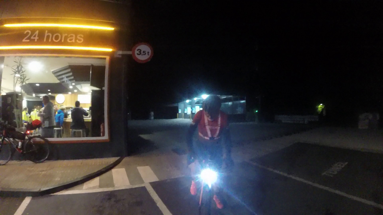 Edgar, on his bike, in a 24h gas station in Lalin at night. He is looking down and his front light is on. Some randonneurs are still inside the gas station bar, which has a neon light that says '24 horas'