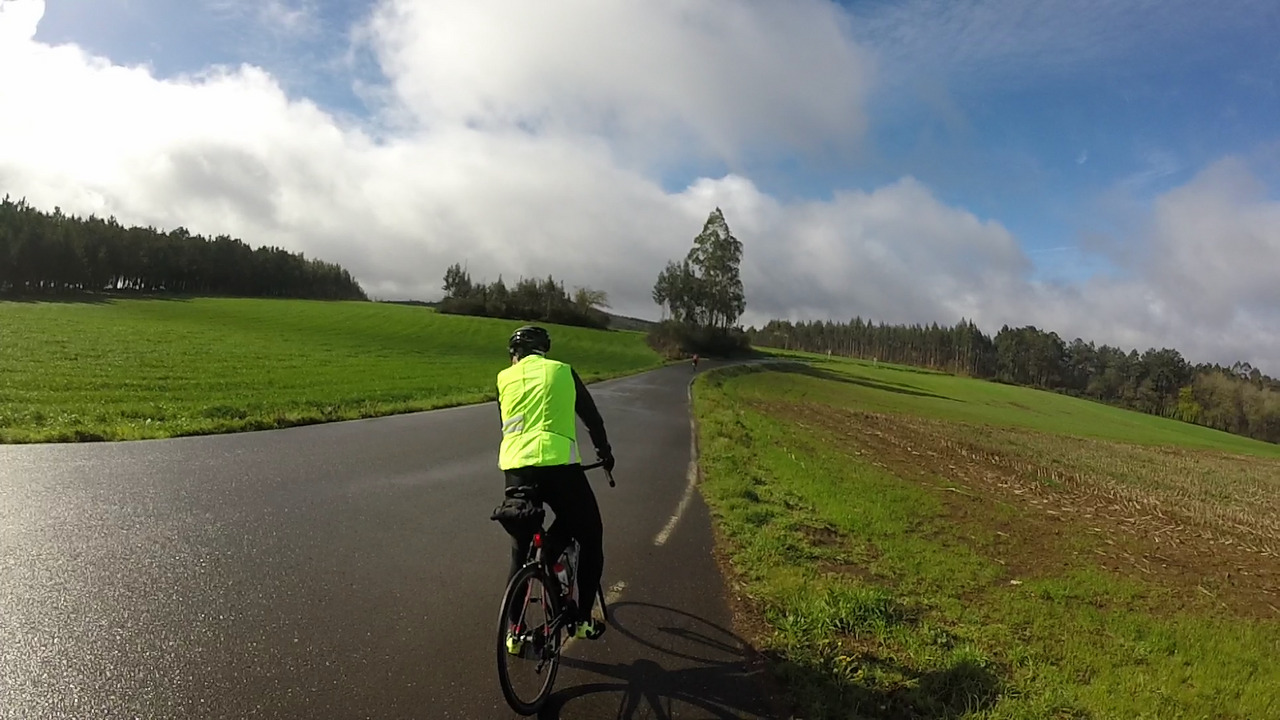 Dani about to resume riding after the stop at the crossroads, edgar already left and can be seen on the road as a small dot. Big clouds cover the blue sky and there are green fields and trees all around the road