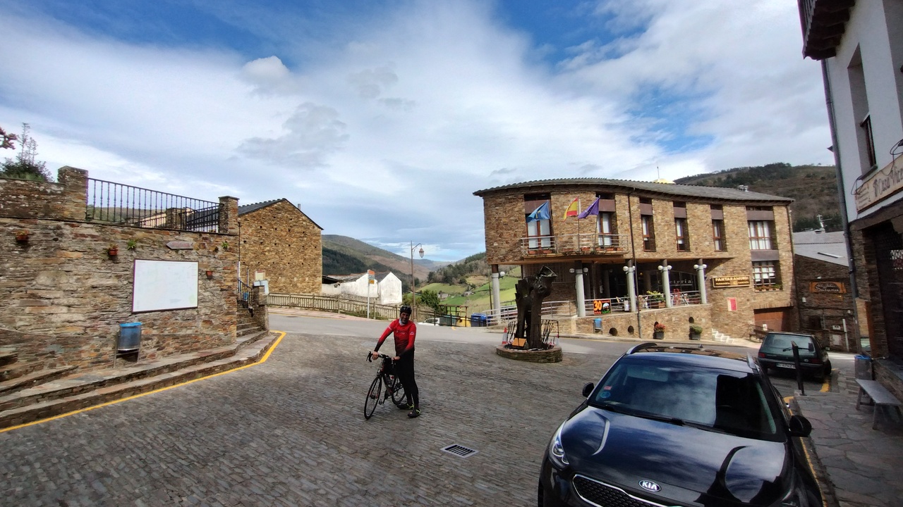 Dani, by his bike, with his right hand made into a fist and pointing up. He is in the middle of the crossroads at the entrance of Taramundi, with several old-style buildings around him.