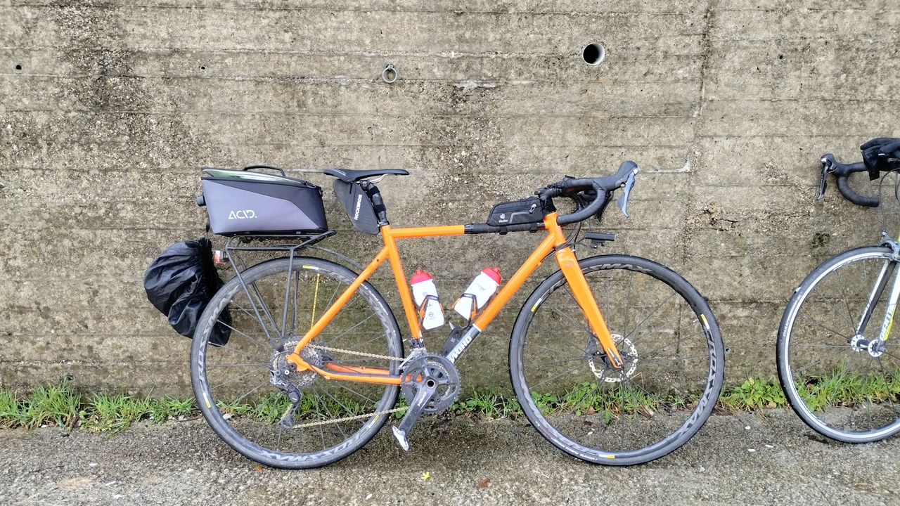 Edgar's woho steel gravel bike, featuring a rear pannier and a huuuge panier bag, mavic crossmax wheels and shimano GRX drivetrain. A beautiful orange and black bike leaning against a concrete wall