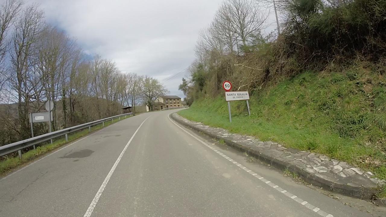 The entrance of Santa Eulalia de Oscos, the road turns right just before an old stone house. Old trees are aligned with the road on the left. On the right there is the sign that marks the start/entrance of the village.