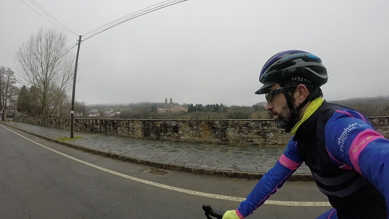 Me from the left side, riding the bike while entering Sobrado dos Monxes, with the monastery towers on the background