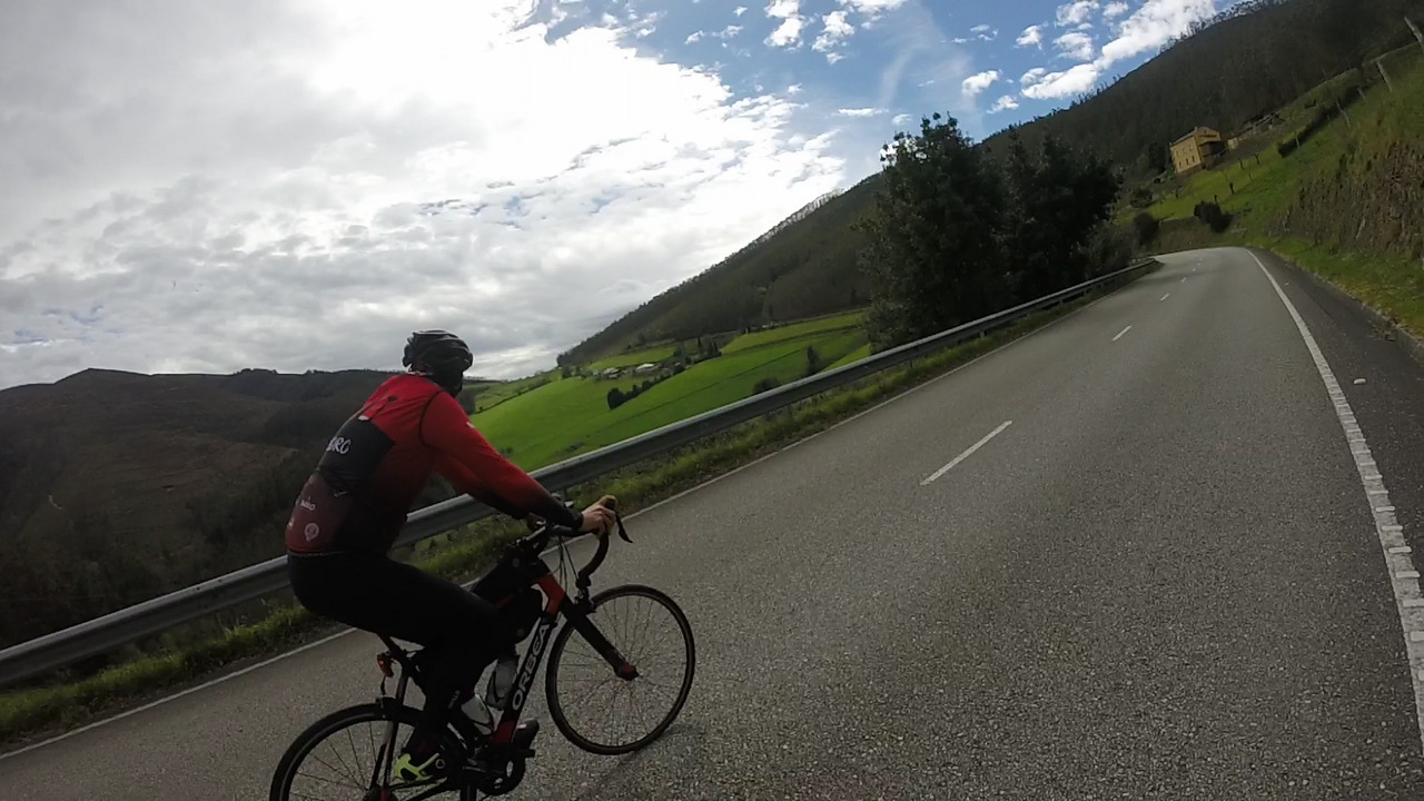Dani is going up on a road that is turning left, there is a nice open view of the valley to his left, with green grass fields and a few hills and small mountains surrounding it. The photo is a bit tilted to the left
