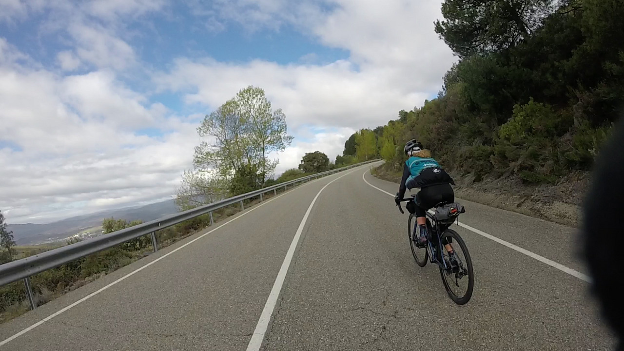 Estefania pushing it up the climb. The road goes up and turns a bit to the right. There are big trees on the right and an open view of the cloudy sky on the left. The photo is a bit tilted to the left