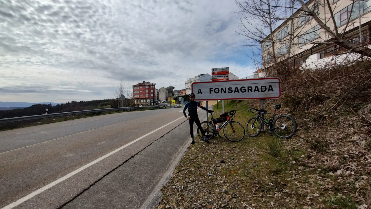Me, by the sign with big letters 'Fonsagrada' at the exit of the village. Both bikes are leaning against the sign and Fonsagrada can be seen on the back