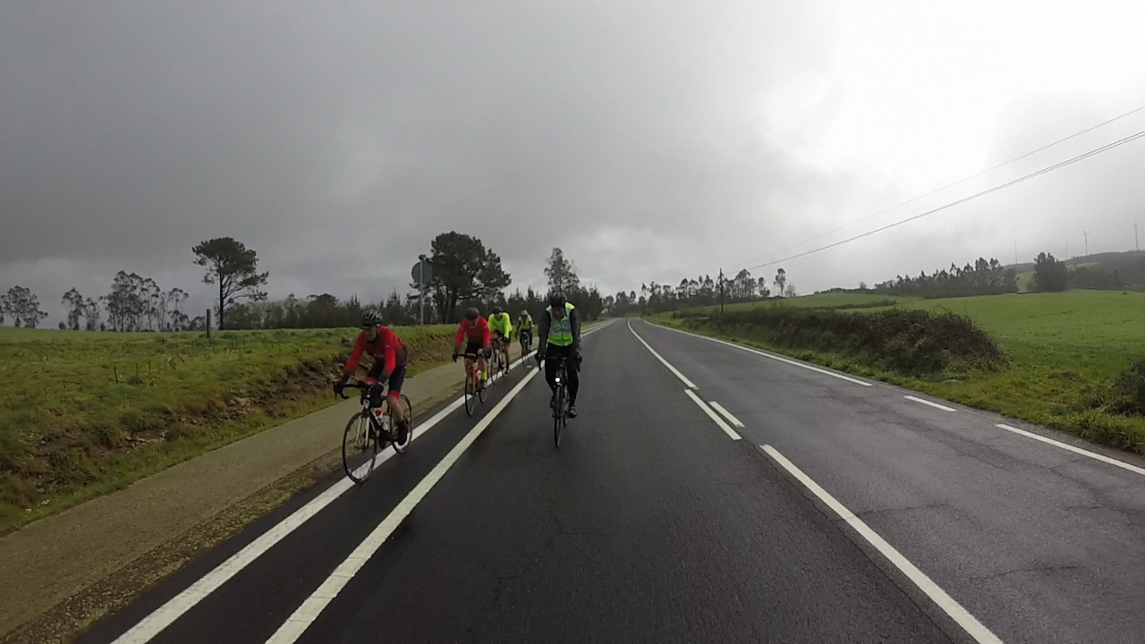 The whole group, except me and Alex, starting to go down another downhill. Road still wet, dark clouds covering the sky except the top-right part of the picture, where it seems like the sun wants to push through the clouds