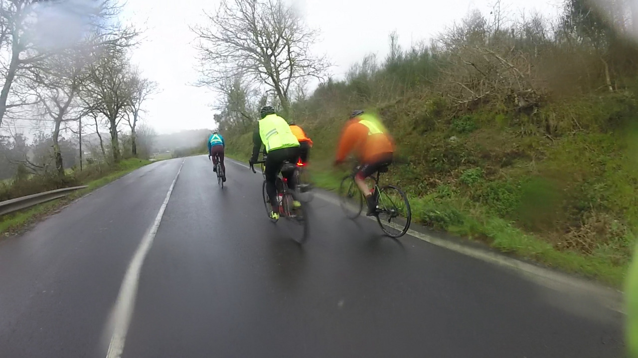 Dani, Jose, Edgar and Alex riding on a wet road towards Zas, trees on both sides of the road, it is raining more now, so the camera is full of rain drops