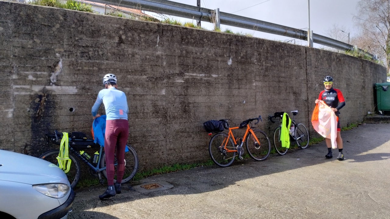 Alex and Oscar removing some layers and hanging them for drying, with their bikes leaning against a concrete wall (wider photo)