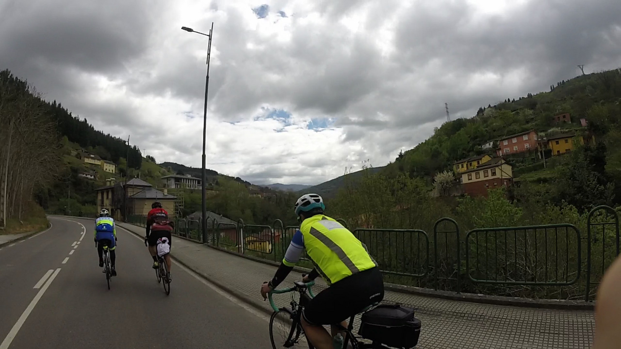 Manolo, Dani and Ferreiro on the road leaving Cangas de Narcea, there are small groups of houses on the hills on both sides, surrounded by trees. The sky is full of dark grey clouds