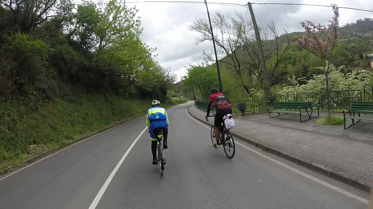 Manolo and Dani, riding side by side on the road leaving Cangas de Narcea. On the right of the photo there are a couple of old benches painted in green and grass and small trees can be seen on both sides of the road