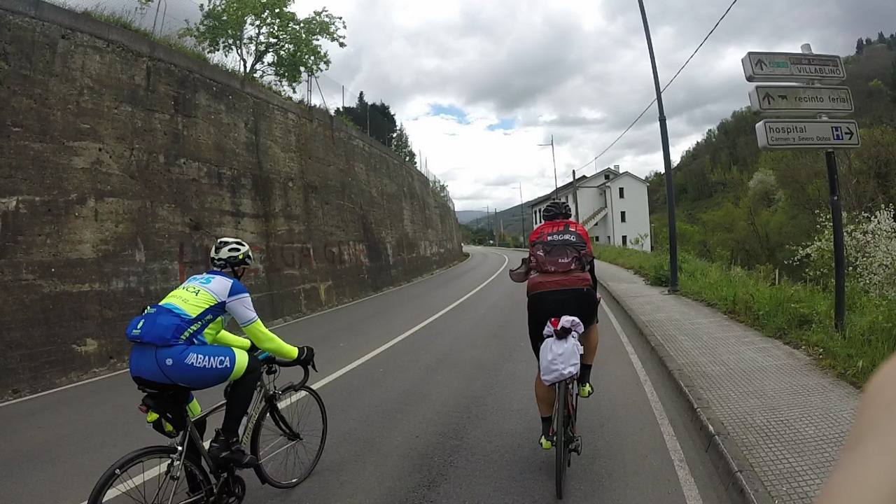 Manolo and Dani, riding on the road leaving Cangas. A tall, grey, cement wall is on their left. On the right there is a sign with arrows pointing to Villablino, the livestock market and the hospital