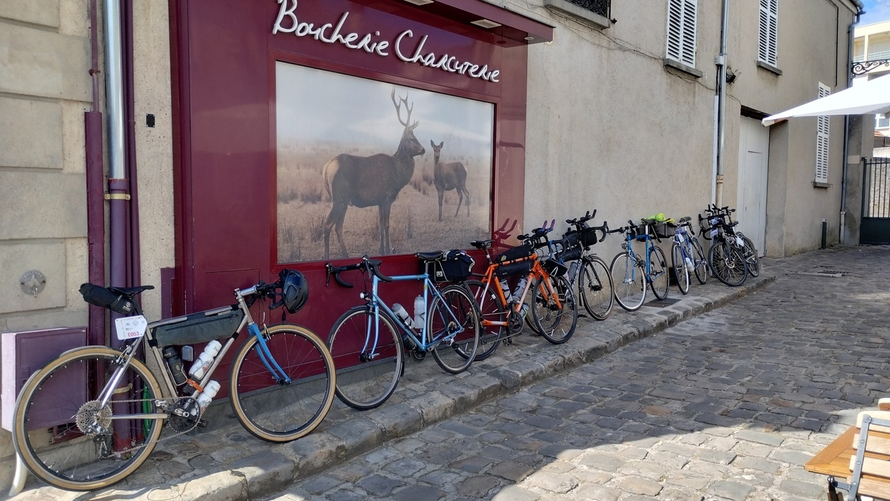 A line of bikes of different frames, materials, wheels and equipment, outside a 'Boucherie' (a butcher)
