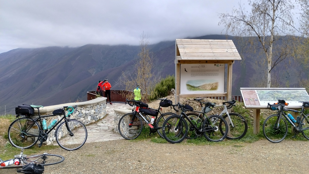 Several road bikes leaning on the signs at 'Mirador da Corza'. There are some mountains at the back of the photo, and a few cyclists enjoying the view from a platform. The sky is full of grey clouds
