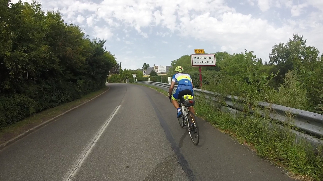 Manolo, standing on his bike, pedaling by the road sign 'Mortagne au Perche' at the entrance of the village