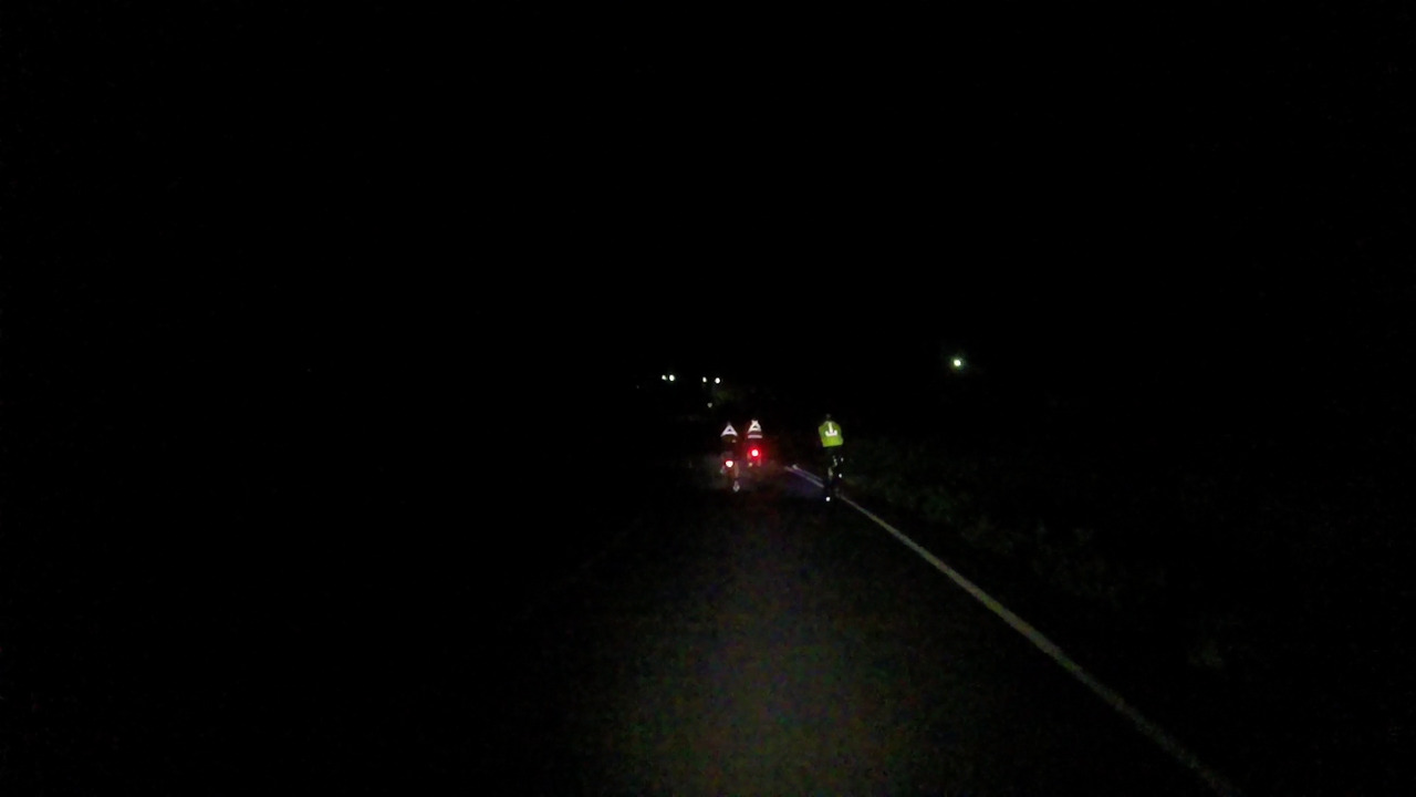 Three randonneurs riding in the middle of a pitch black night, everything around them is black, my front light make them visible due to their reflective clothes, their tail lights are bright red dots