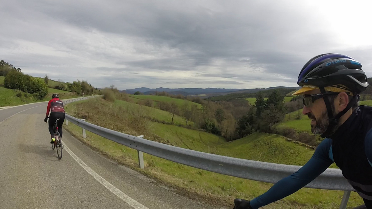 Dani on the left, me on the right, approaching a turn to the right on the road at the start of another downhill section. To the right there is yet another open view of the valley with its green fields. The sky is full of light grey clouds.