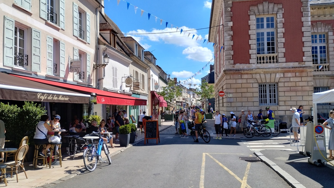 One of the streets in Rambouillet, quite crowded, lots of people, cyclists... On the left there are some cafés with people sitting at tables in the terraces