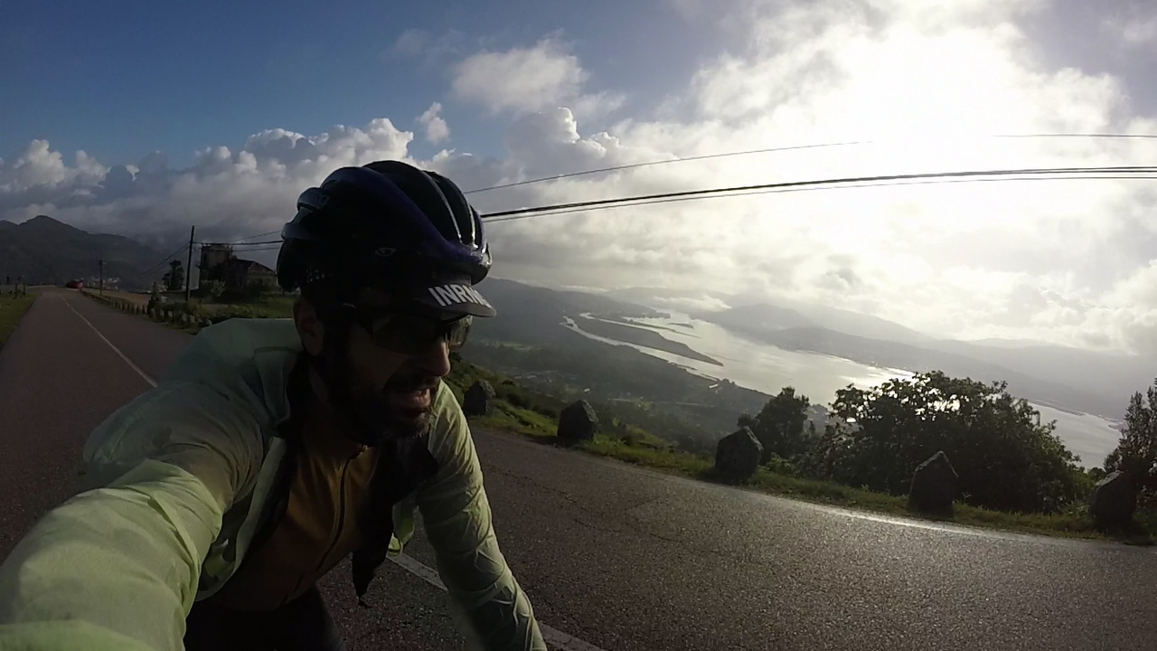 Me, riding up to the top of Santa Tegra, behind me is a gorgeous view of the Miño river. The sun is shining from behind some white clouds, which makes the sky look all white