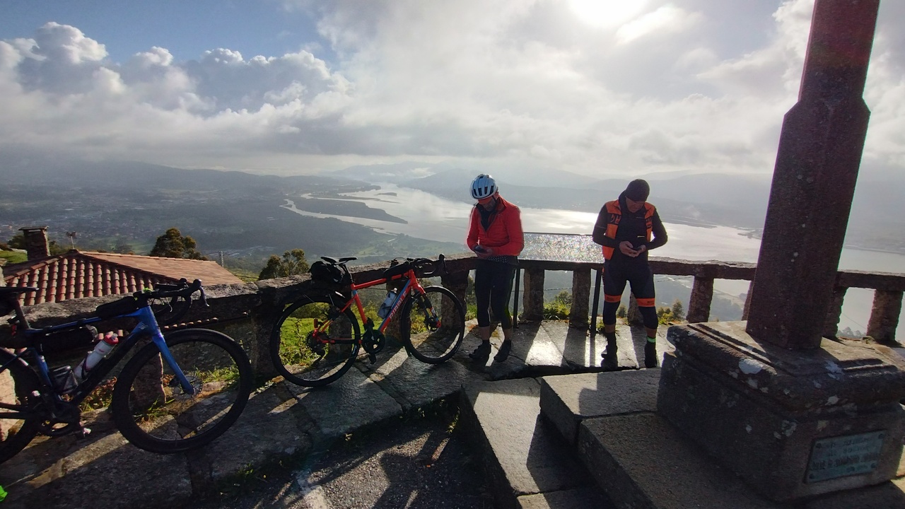 Graeme and Salva at the top of Santa Trega, both looking at their phones, with a gorgeous view of the Miño behind them