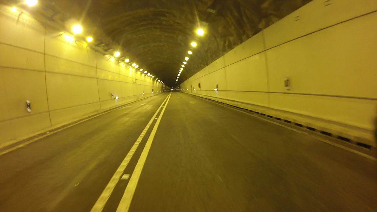 The inside of the tunnel, with yellow lights on a dark ceiling and big white walls on both sides. A double white straight line separates the two lanes of the road passing through the tunnel